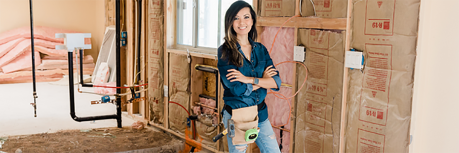 A lady in jeans, a blue shirt, and tool belt stands smiling in a building under construction. Exposed insulations and pipes are visible behind her. 