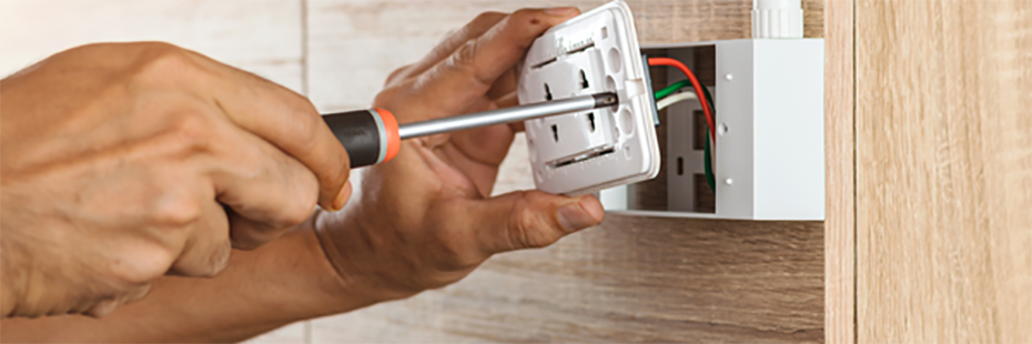 A man screwing the front panel of an electrical outlet onto the body of the outlet. Red, green, and white wires can be seen within the outlet. The white, plastic outlet is mounted on a wooden wall. 