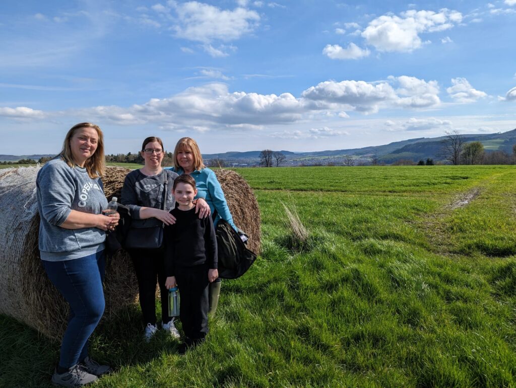 From left to right: Charlotte, Helen, Noah, and Carole stand in front of a large, round hay bail in a grassy field. They are positioned to the left of the image. 