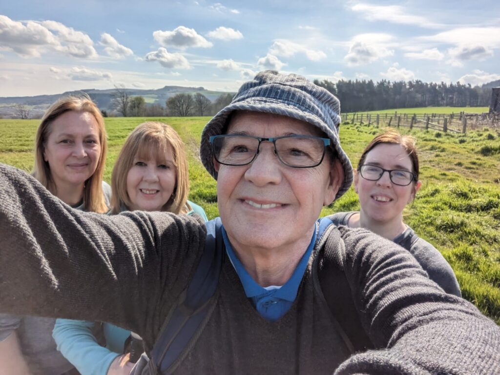 Tony's head and torso are in the foreground as he is taking a selfie of himself and the rest of the group stood behind him (from left to right: Charlotte, Carole, and Helen). The group are in a grassy field. Green hills and tall trees can be seen in the background. 