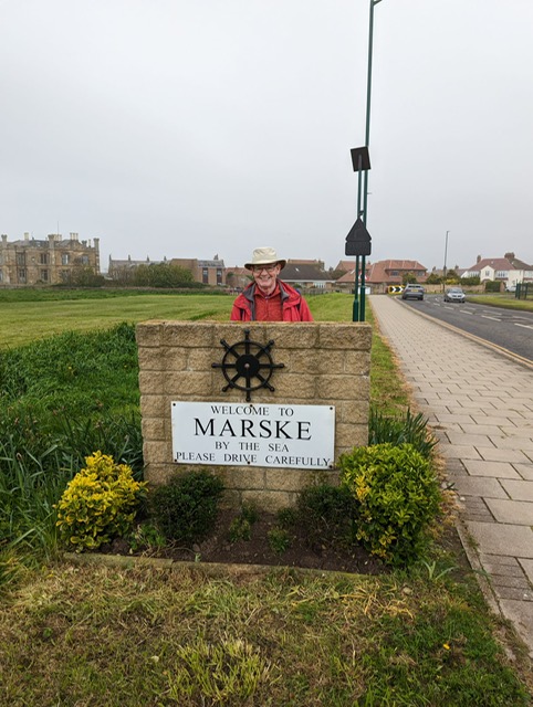 Tony, an older mad in a white hat and red raincoat, stands behind a small stone wall surrounded by grass and flowers. On the wall is a sign that reads "Welcome to Maske By The Sea Please Drive Carefully". 