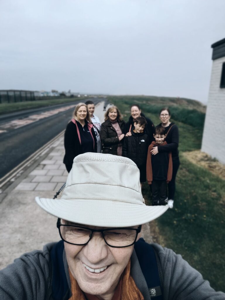 Tony's head and torso are in the foreground as he is taking a selfie of himself and the rest of the group stood behind him (from left to right: Charlotte, Sue, Carole, Sally, Harry, Helen and Noah). The group stood on a stone pathway. To the left of them is a road and to the right of them are the grassy tops of cliffs. The sky is grey but everyone is smiling. 