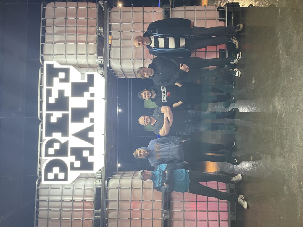 The Dispatch Team are stood side-by-side in a line facing the camera. Behind and above them is a large LED sign that reads "Drift Hall". The font is pixelated in the style of '80s computer games.   