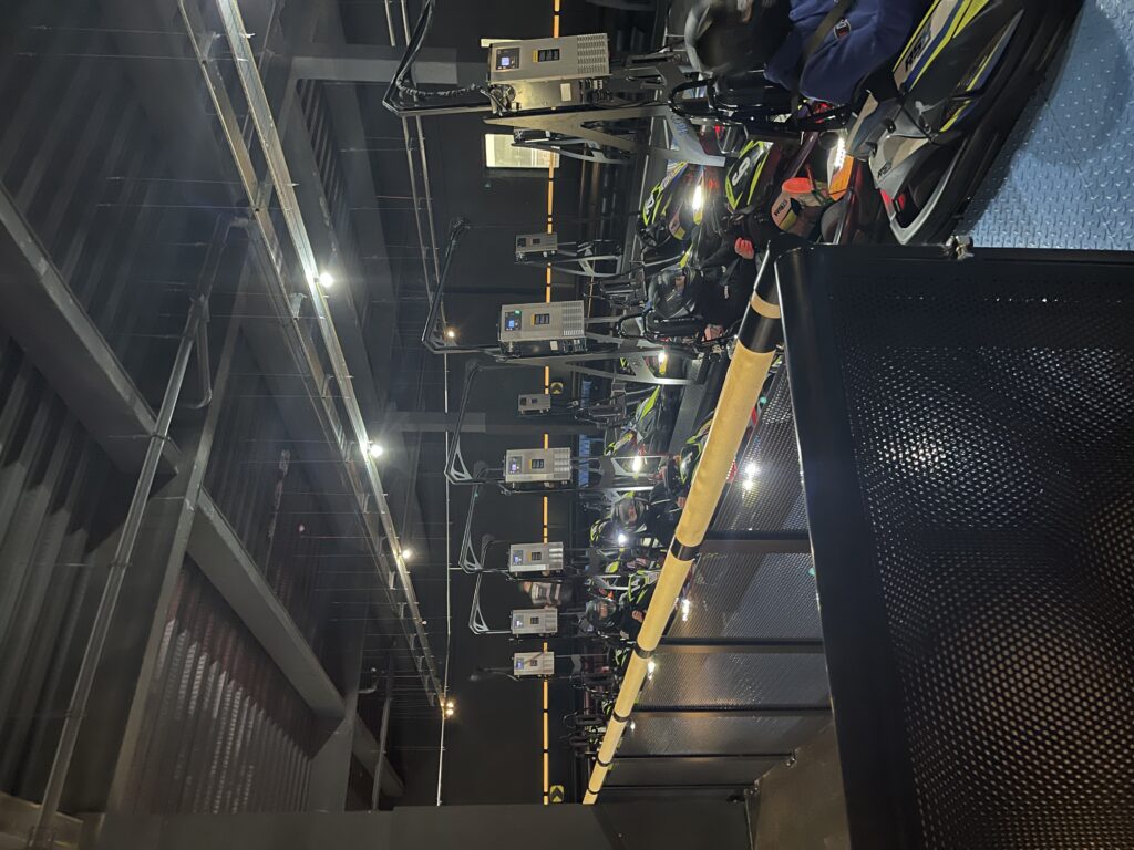 Black electric go-karts with neon yellow go-faster stripes line up on the indoor track awaiting the start of the race. The drivers face forwards and are wearing black motorbike-style helmets. 