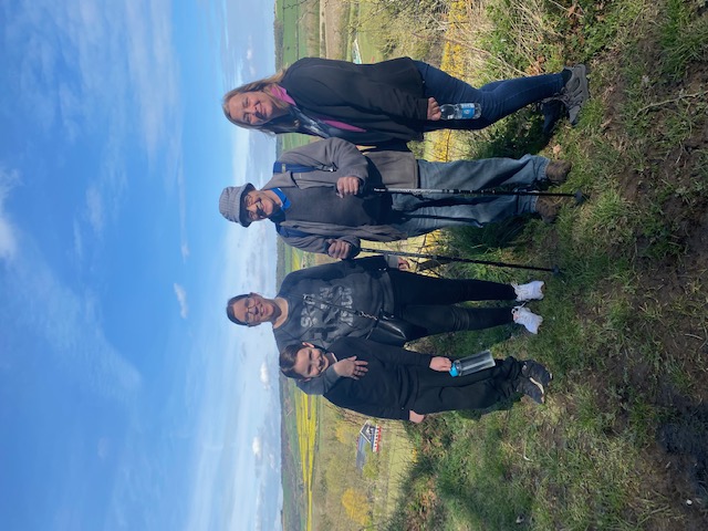 From left to right Noah (a small boy dressed on black), Helen (Noah's mother; she stands with an arm around his shoulders), Tony (an older man wearing a hat, blue rucksack and holding walking sticks), and Charlotte (a woman dressed in jeans and black waterproof jacket) stand on a grassy bank. They smile at the camera. Behind them are rolling fields of green of yellow broken up by rows of hedges and trees. 