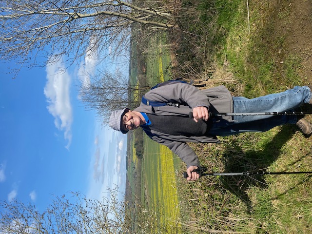 Tony stands smiling at the camera. He is wearing a grey hat, fleece and blue jeans. A blue rucksack is on his back and he is holding two walking sticks - one in each hand. He stands on a grassy path framed by silver birch trees that have just begun to bloom. Behind him green and yellow fields stretch all the way to the horizon. 