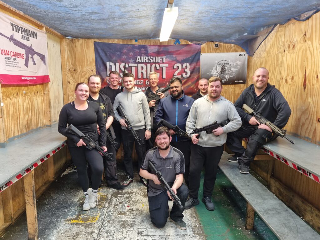 The Calibration team gather in a room with wooden walls and grey benches on either side. They are looking at the camera smiling. They are all holding a BB gun. One team member, Adam, kneels in front of the rest of the department. 