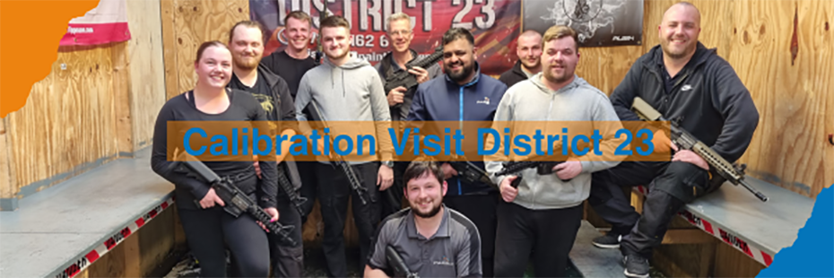 The Calibration team gather in a room with wooden walls and grey benches on either side. They are looking at the camera smiling. They are all holding a BB gun. One team member, Adam, kneels in front of the rest of the department. In the centre, large blue text on a translucent orange background reads "Calibration Visit District 23".  