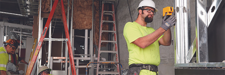 On the right of the image a workman in a lime green t-shirt and white hard hat is holding a Fluke laser level and pointing it at the metal beams in front of him. Behind him, the work site is littered with ladders and tools, and two other work men in hard hats and high vis are completing tasks.