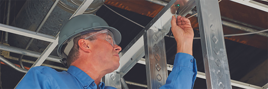 A man in a grey hardhat, blue shirt, and protective glasses is marking a metal beam where the green dot from a Fluke point laser has been projected.