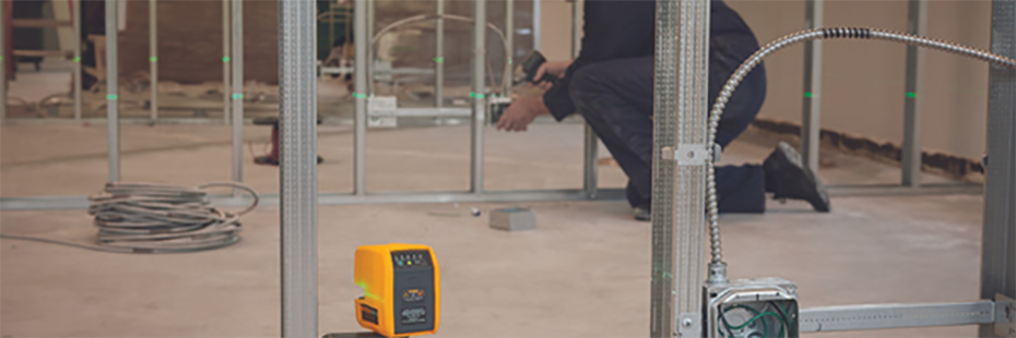 At the bottom centre of the image a Fluke Laser Level is projecting a green, horizontal laser line through the work site dotted with metal beams. Two large coils of electrical cabling lie on the concrete floor while a man in blue overalls carries out his task in the background.