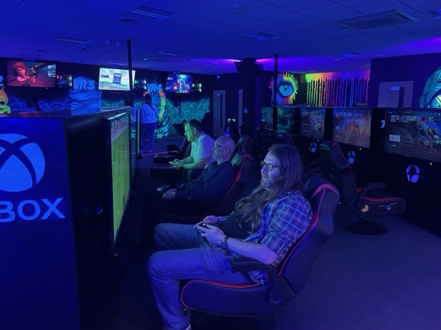Three men sit in comfy gaming chairs in a dark room with neon lighting and decorated with graffiti-esque murals painted in glow-in-the-dark paint. The men are arranged in a line going away from the camera; from front to back: Stacey, Marc, Mat. In front of them are flat-screen TVs. All are playing FIFA.
