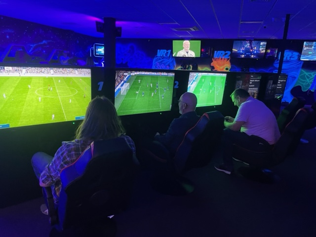 Three men sit in a row with their backs to the camera. The men are arranged from left to right: Stacey, Marc, Mat. They are in a dark room illuminated by neon light and decorated with fluorescent paint. All are sat in comfy gaming chairs and playing FIFA on flat screen TVs. 