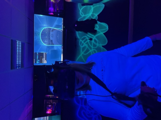 Rachael, a young woman with shoulder-length hair, stands in the middle of a neon illuminated dark room. She is wearing a VR headset and behind and above her is a flat screen TV showing a scene from the VR game. 