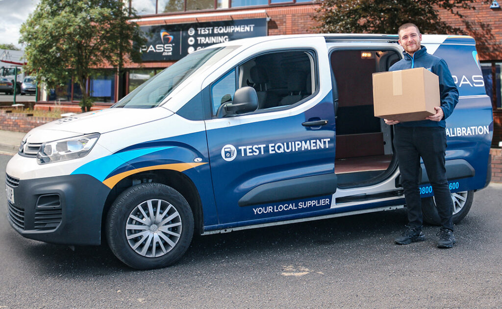 Bradley, a young man with short brown hair, is wearing a PASS Ltd blue jacket. He is standing in front of a PASS van holding a box. The van is parked on a diagonal in front of the PASS Ltd's headquarters in Stockton. A big, blue sign with the PASS logo on identifies the red brick building as PASS' offices. Two green, leafy trees from the image. 