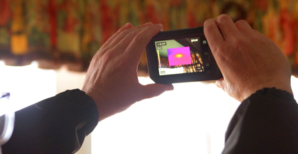 A man holds the Teledyne FLIR C5 Thermal. He is inside pointing it at the ceiling, trying to locate a hive internally as part of a bee removal mission. Picture-in-Picture mode is engaged and on the screen is a small thermal image revealing the location of a hive. It sits in the centre of a digital image of the ceiling.  