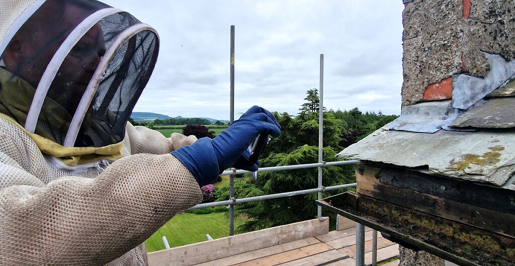 A man in a bee suit holds the Teledyne FLIR C5 Thermal Camera in gloved hands. He is standing on scaffolding, pointing the camera at roof guttering. He is using thermal imaging to locate a hive from the outside as part of a bee removal mission. The green North Yorkshire countryside rolls out behind him. 