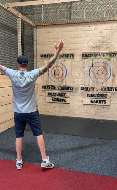Lee, a man in a grey t-shirt and navy shorts and cap, stands victorious with his arms up in the air. His axe is lodged near the centre of the wooden target. 