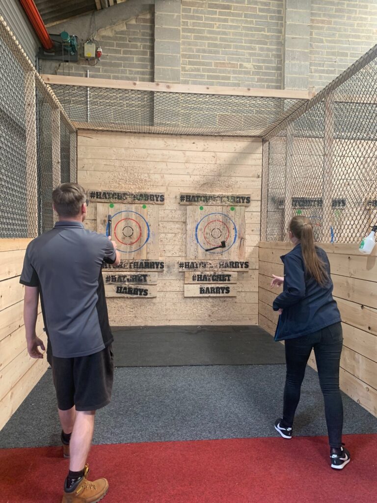 Arturs, a man in a grey t-shirt and shorts stands on the left having thrown his axe at the wooden target where it has lodged successful. To his right, Stevie, a lady in black jeans and a navy jacket with brown hair pulled back into a ponytail watches as the axe she has just thrown bounces off the wooden target. 