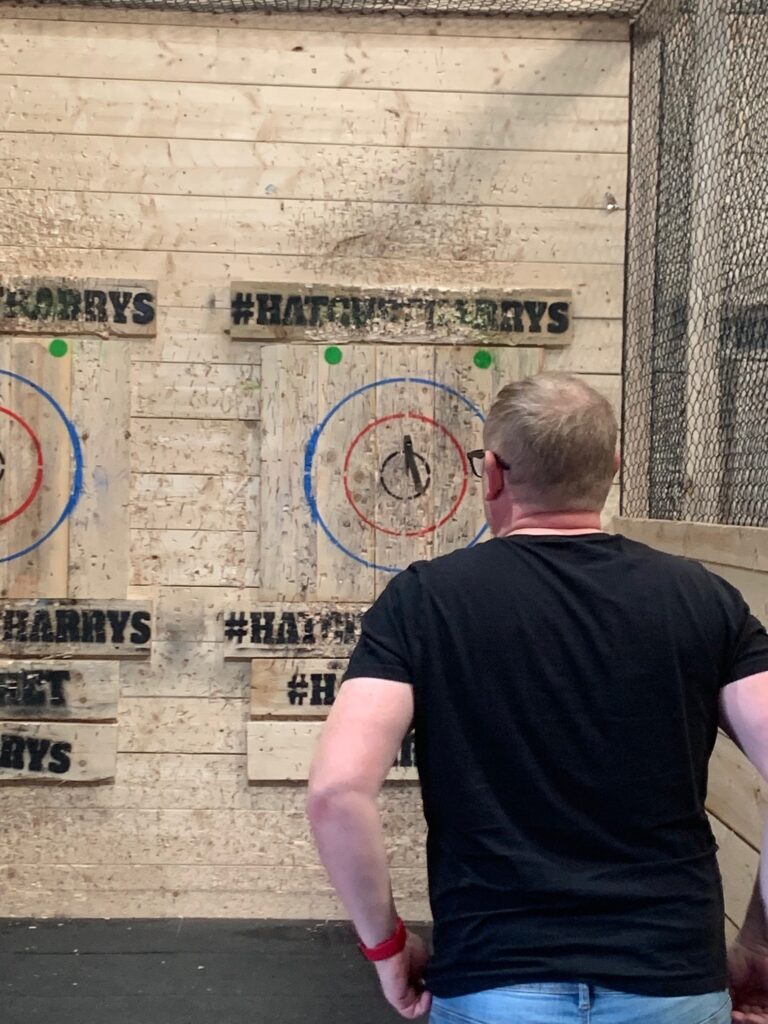 Simon, a man with ginger hair, a black t-shirt, and blue jeans, walks towards the wooden target to retrieve his axe which has struck the centre. 