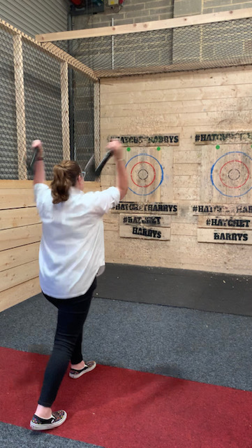 Sally, a lady in a long white shirt and jeans with brown hair pulled back into a short ponytail, stands with her feet in parallel, one leg in front. Her arms are up and behind her head, ready to throw the axes she holds in each hand at the wooden target. 
