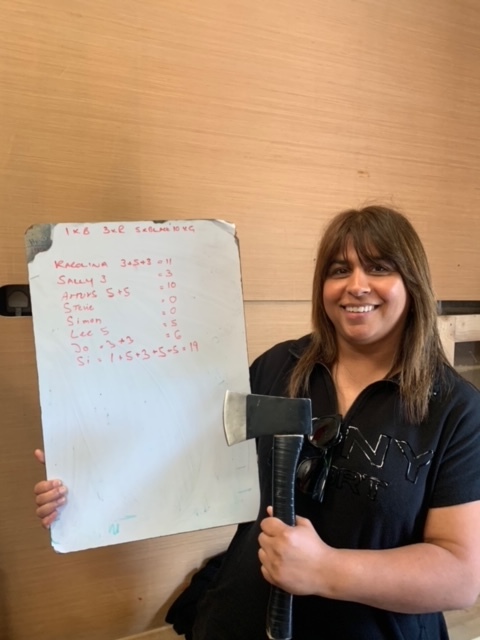 Si, a lady dressed all in black with shoulder-length brown hair, stands in front of a wooden wall. She is smiling and holding an axe in her left hand a white board in her left. The whiteboard lists everyone's scores, Si has the most points with 19. 