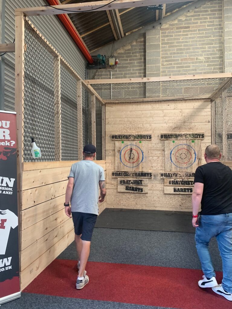 Lee (left), a man in a grey t-shirt and navy shorts and cap, and Simon (right), a man with ginger hair, a black t-shirt, and blue jeans, walk towards the wooden targets to retrieve their axes.  