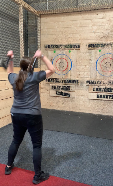 Jo, a lady in a grey t-shirt and jeans with brown hair pulled back into a low ponytail, stands with her feet in wide. Her arms are up and behind her head, ready to throw the axes she holds in each hand at the wooden target. 
