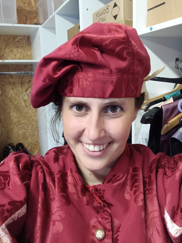 Bethany, a lady with dark hair and brown eyes, is dressed in a red medieval costume with a floppy red hat. She is taking a smiling selfie. 