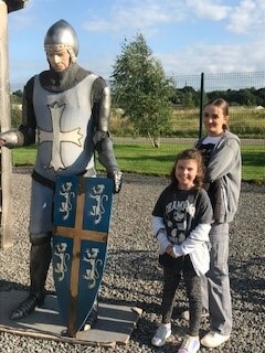 On the left is a medieval knight dressed in armour. A blue shield with a yellow cross and four, white, rearing, roaring lions. On the right, two young girls with brown hair are smiling.  