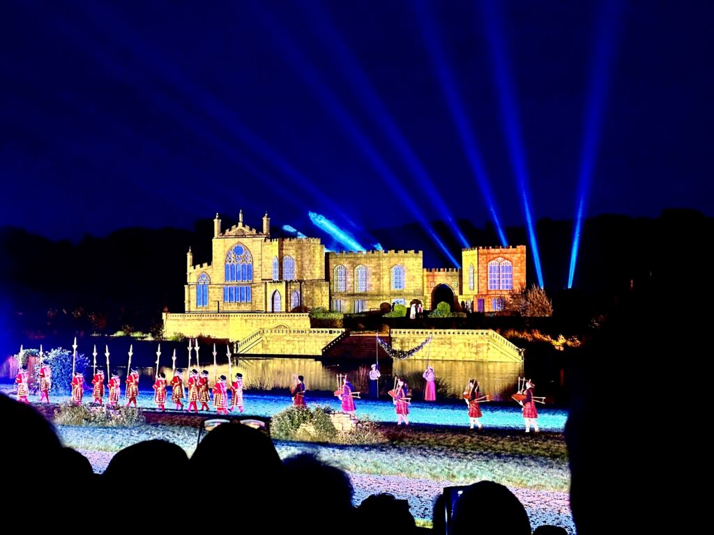Framed by a navy night sky. People holding spears or bagpipes and dressed as beefeaters but without bearskin hats are marching across a field in front of a river. On the opposite bank is a stone castle with blue lights shooting out behind it. 