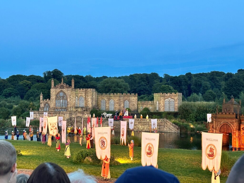 At dusk, people in medieval dress are holding medieval banners. They stand on a neatly cut lawn in front of a wide river. On the opposite bank is a small stone castle framed by trees.  