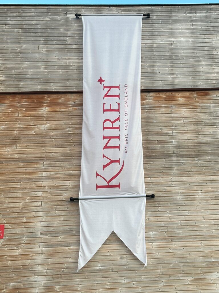 A white medieval-style banner hangs vertically on a wooden wall. Red lettering, written vertically up the banner reads "Kynren". 
