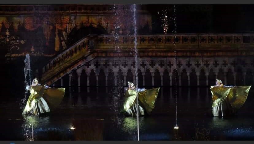 Three people wearing gold dresses with gold fanning sleeves. They stand in a line, all looking and pointing both arms to the right. They are stood in a fountain/pool with water spurting up around them. In the background is a stone castle. 