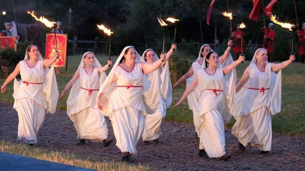 Seven women in white, floaty dresses with a red trimmed collar, red rope belt, and white headdress, hold fire torches above their heads as they march in pairs down a sand path. Behind them, men dressed as knights line the edges of the fields. 