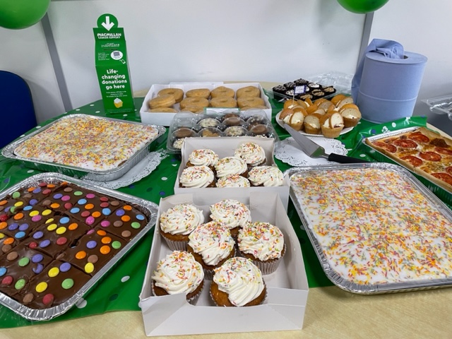 A table full of baked goods including iced traybakes, frosted cupcakes, and a tomato quiche. The baked goods sit on a green spotted table cloth. At the back of the table is a green donation box. 
