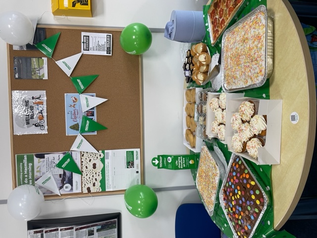 A round table flush against the wall. On the table is a green spotted tablecloth and on top of this are a selection of traybakes, cupcakes, doughnuts, and a quiche. Behind the table is a noticeboard draped in Macmillan green and white bunting and framed by Macmillan balloons in each corner.  