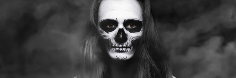 Black and white image of a woman with her face painted like a skeleton. She stands in a smokey room, looking straight at the camera. The only colour in the image is her brown eyes. 