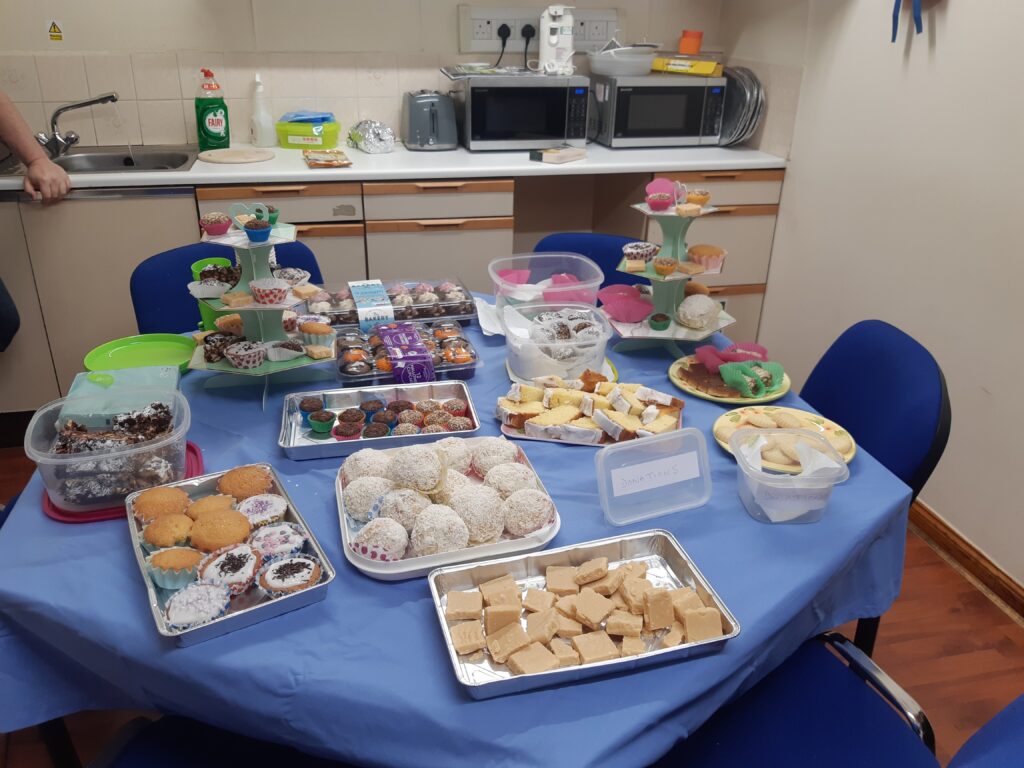 In an office kitchen, a pentagonal table shrouded in a blue tablecloth stands in the centre and is surrounded by blue chairs. On the table are an array of baked goods including traybakes, cupcakes, brownies, and blondies. 
