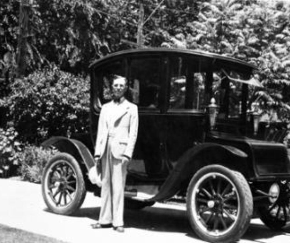 An older Oliver Fritcle wearing a white suit and bowtie stands in front of a large, black, closed-carriage electric car.  In the background are tall, fir trees. 