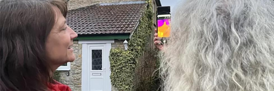 Two women use a smartphone with a thermal imaging attachment to inspect a stone house with a white door and climbing plant along one wall. 