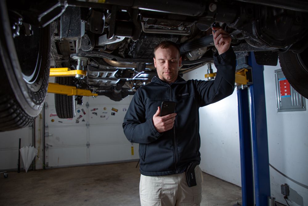 A mechanic stands under a raised vehicle. He is using the FLIR One Edge Pro to inspect the components of the vehicle while viewing the thermal images on his smartphone. 