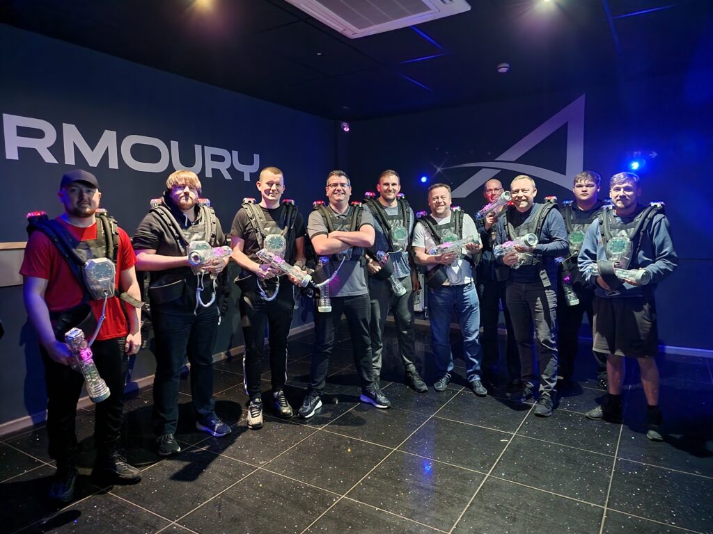Dressed in harnesses and holding sci-fi style, transparent guns, the Sales team stand in a semi-circle smiling at the camera.  