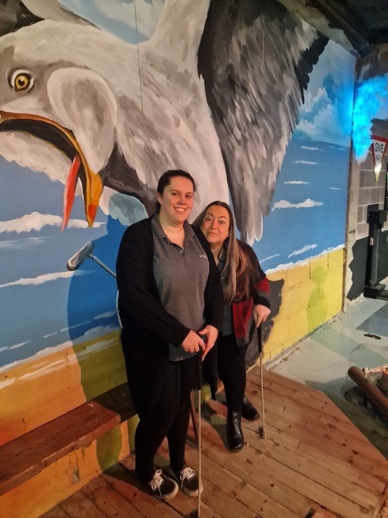 Collette and Clare stand with mini-golf clubs in front of a wall with a mural of a flying giant seagull with a gaping mouth/beak. 