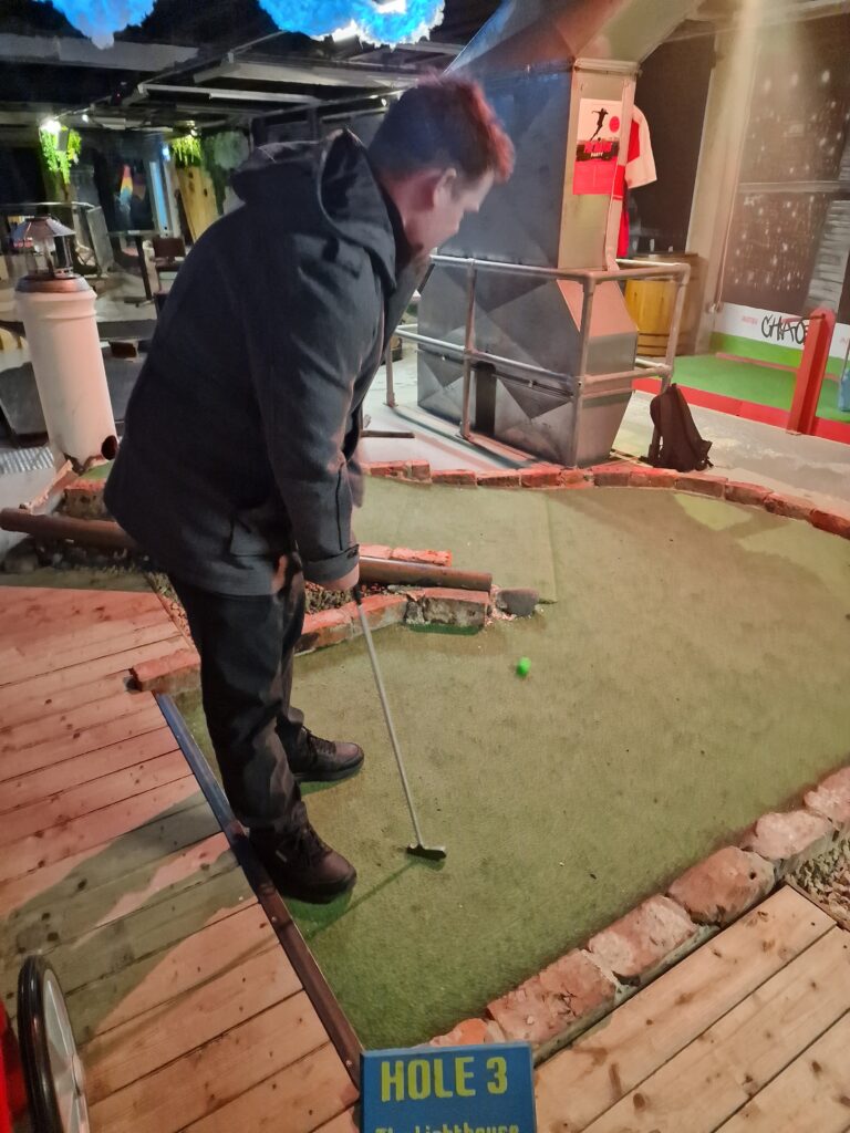 Michael stands on a crazy/mini golf putting green. Despite the course being inside, he is wearing a thick, dark winter coat with a hood. He is in the middle of putting a neon green golf ball. 