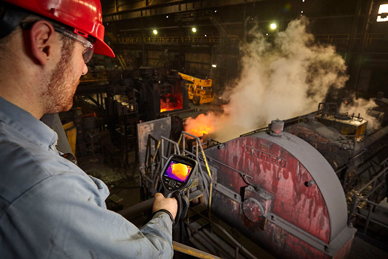 A man in a red hardhat uses the FLIR E76 to inspect a mechanical furnace.