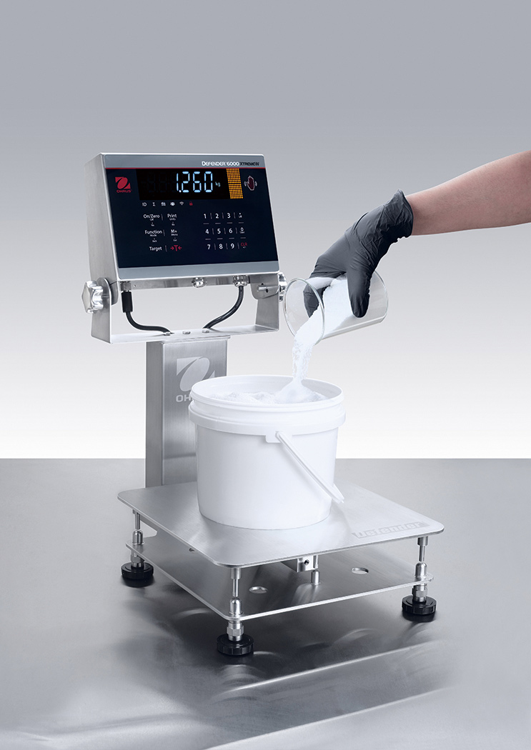 Ohaus Defender 6000 Stainless Steel IP68/IP69K Washdown i-D61XW Bench Scales with a bucket of powder on top.