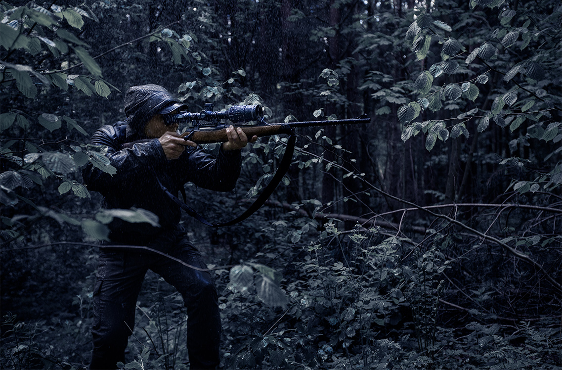 A hunter aiming at a tartget through the scope while camoflauged in the greenery.