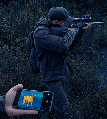 A hunter taking aim at their prey through the scope which is displayed on a mobile phone held by their hunting buddy.