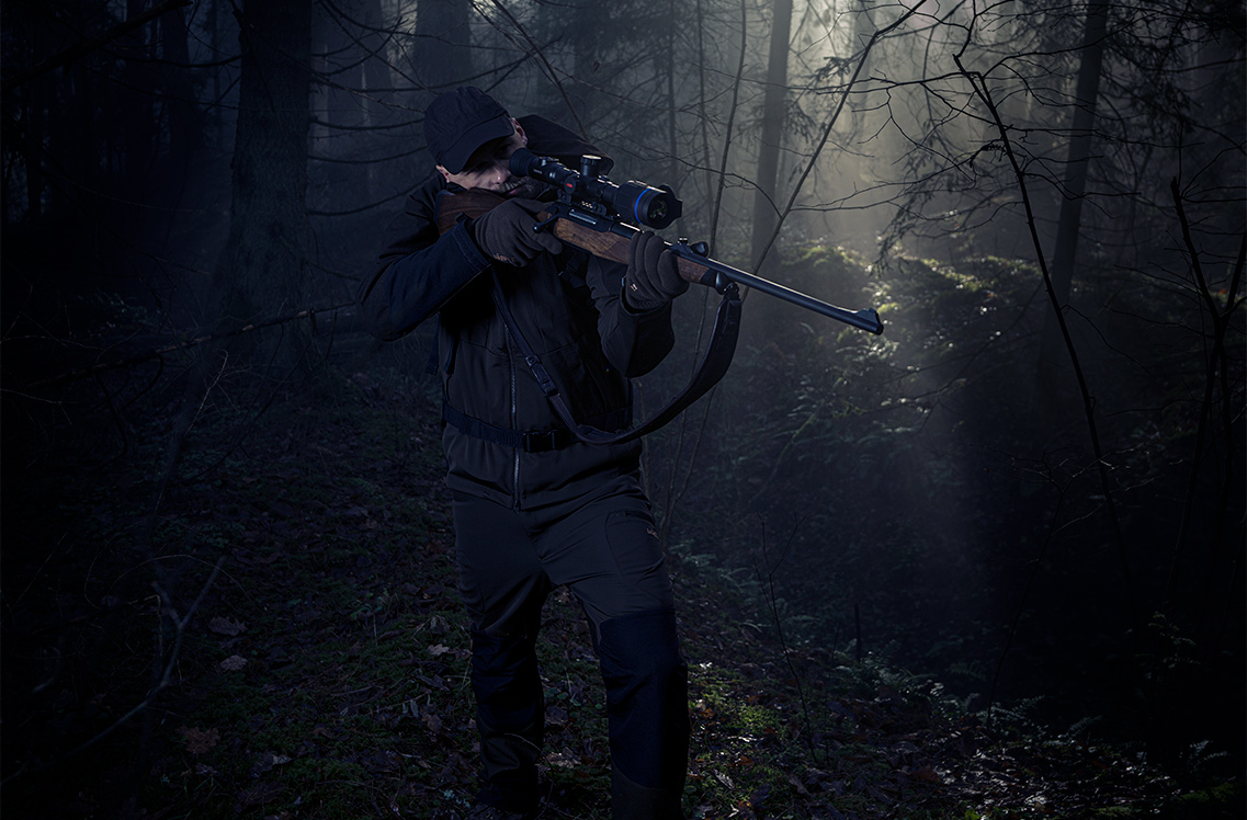 A hunter aiming at a target through the scope while in a wooded area.